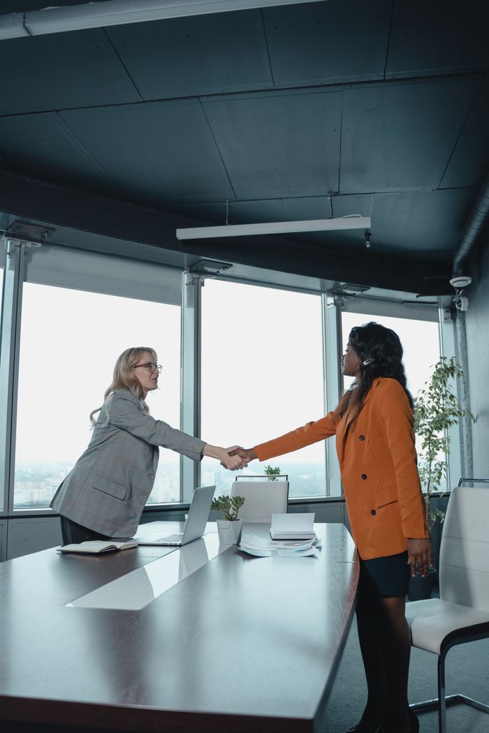 Two businesswomen shake hands across a conference table in a modern office setting global talent finder