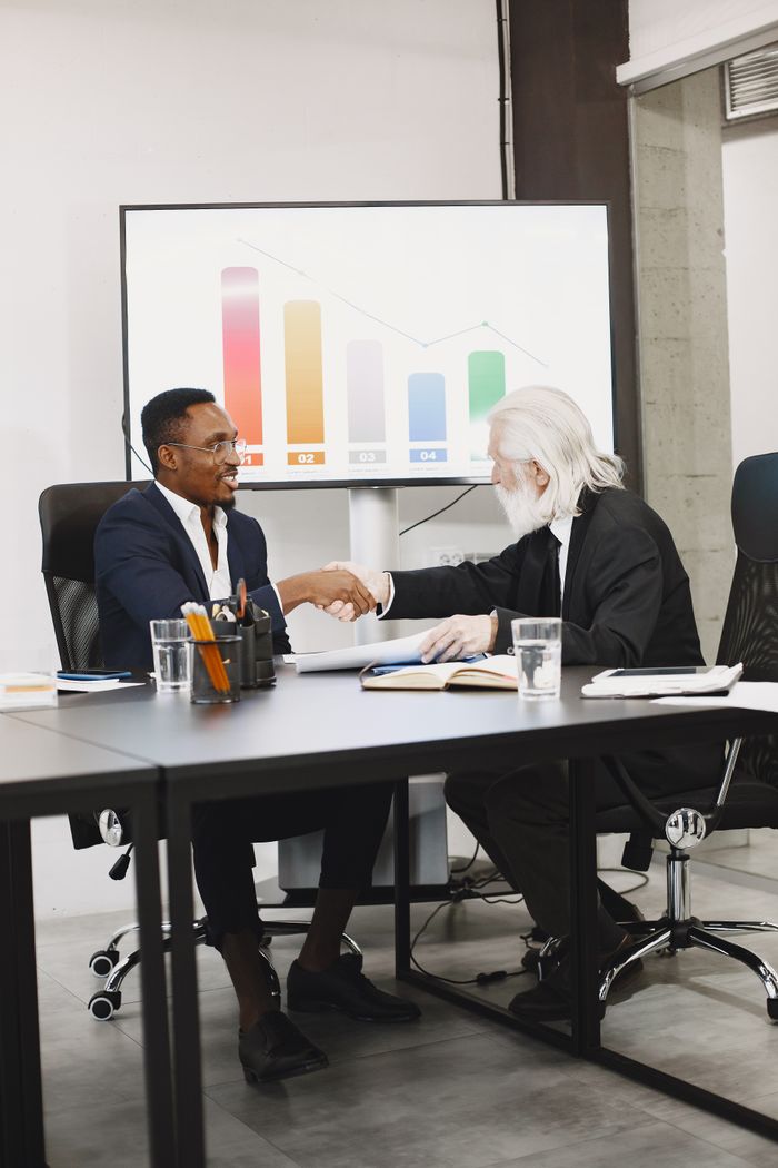 Two diverse businessmen in suits shaking hands over a desk in a modern office global talent finder